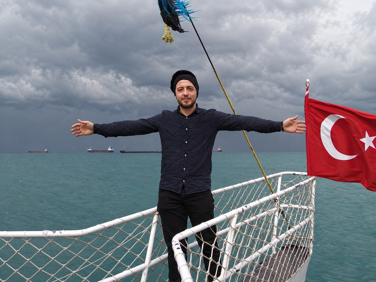 Basel on a boat at sea, arms wide, a Turkish flag beside him.